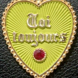 May include: Heart-shaped pendant with a light green background and gold trim. The pendant has the words "Toi toujours" in silver lettering and a red gemstone at the bottom. The pendant is likely made of metal.