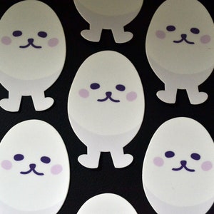 Op de afbeelding: Een verzameling witte, eivormige stickers met paarse ogen en roze wangen. Elke sticker heeft een simpel, cartoonachtig gezicht met een kleine neus en een kleine mond.