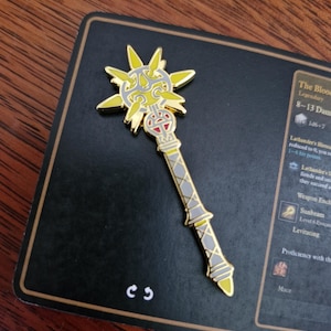 May include: A gold-coloured mace pin featuring a sunburst design at the top and a grey and yellow handle. The pin rests on a black card with text that reads "The Blood Legendary 8-13 Damage".