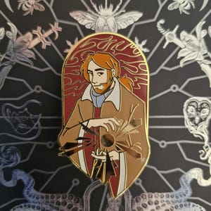 May include: A gold-rimmed enamel pin featuring a stylized illustration of a person with reddish-brown hair and a beard, wearing a tan coat over a red garment, holding a glowing, shattered circular object with sharp, radiating shards.