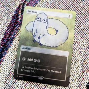 May include: A Sol Ring card featuring a cartoon creature with a ring-shaped body and large eyes. The card has text that reads "Sol Ring" and "Artifact". The card also has the text "It tends to be attracted to the smell of magic ink."