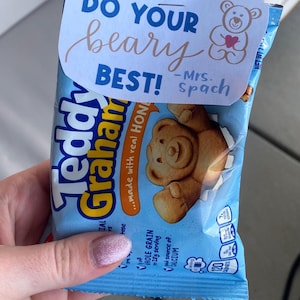 May include: A light blue package of Teddy Grahams, held by a hand with pink glitter nail polish. The package has a white tag that says "Do Your Beary Best!" with a teddy bear graphic and "-Mrs. Spach". The package also says "made with real HON" and "WHOLE GRAIN".