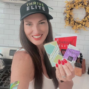 May include: A woman wearing a black baseball cap with the text "THRIVE ELITE" in white letters is holding two Thrive Elite products. The products are a purple and white packet and a green and white box. The woman is wearing a white tank top and has red nail polish on her nails.