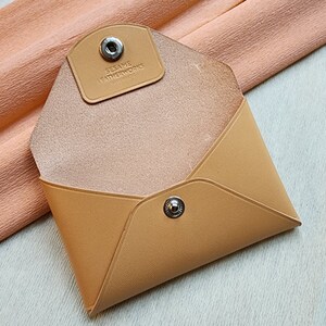 May include: A tan leather envelope-style wallet with a snap closure and the text "SESAME LEATHERWORKS" embossed on the front.