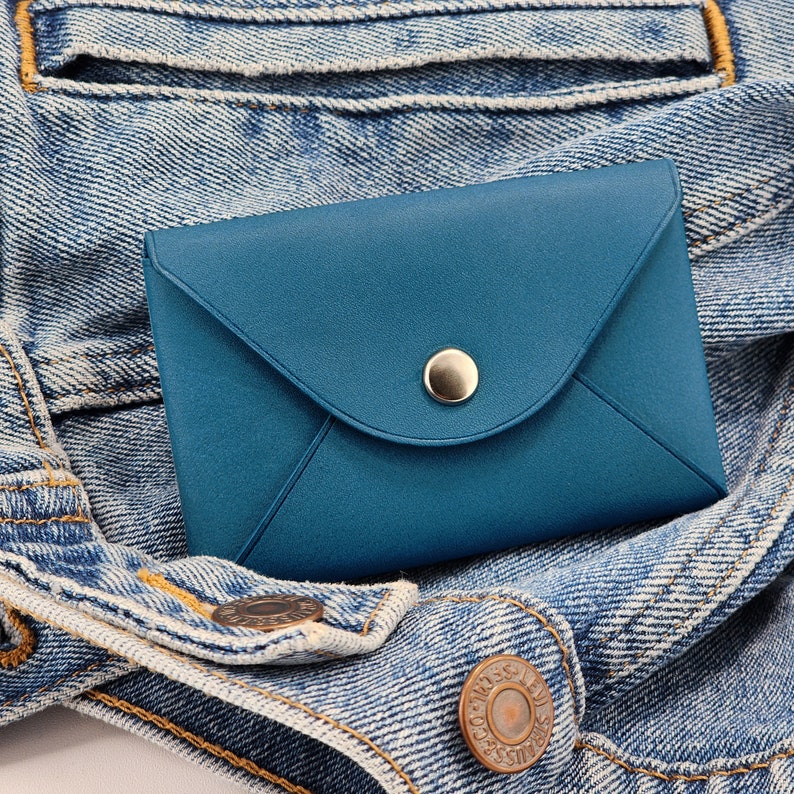 May include: A small, blue leather envelope wallet with a silver button closure. The wallet is sitting on a blue denim jacket.