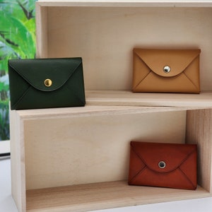 May include: Three leather envelope style wallets displayed on a wooden shelf. The wallets are in green, brown, and red.