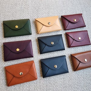 May include: Nine leather card holders in various colors, including green, burgundy, brown, tan, and navy blue. Each holder has a gold button closure and an envelope-style design.