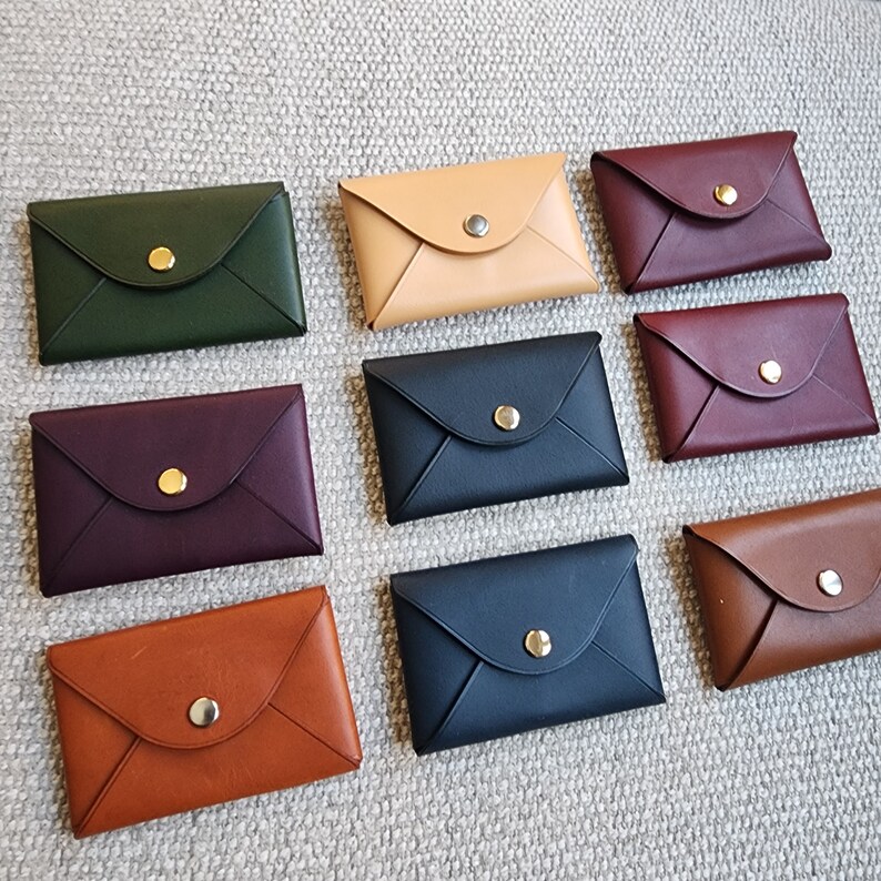 May include: Nine leather card holders in various colors, including green, burgundy, brown, tan, and navy blue. Each holder has a gold button closure and an envelope-style design.
