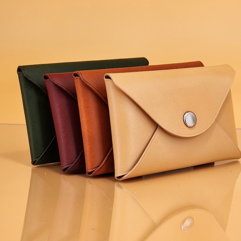 May include: Four leather wallets in different colors, including green, burgundy, brown, and beige. The wallets are arranged in a row, with the beige wallet in the foreground. The beige wallet has a flap closure and a silver button.