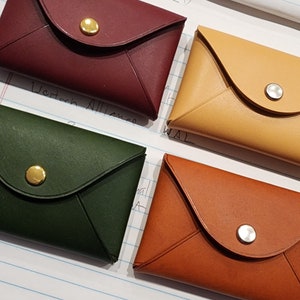 May include: Four leather envelope-style wallets in different colors: burgundy, yellow, green, and brown. Each wallet has a gold or silver button closure.