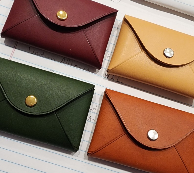 May include: Four leather envelope-style wallets in different colors: burgundy, yellow, green, and brown. Each wallet has a gold or silver button closure.