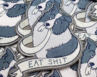 May include: A collection of embroidered patches featuring a stylized illustration of a border collie dog with dark gray and white fur. The dog is depicted holding a white banner in its mouth that has the text EAT SHIT embroidered in dark gray thread.