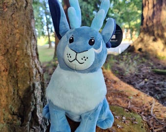 May include: A plush blue creature with large ears and antlers. The stuffed animal has a light blue chest and face with a black stitched nose and mouth. The toy is sitting on a tree stump in a natural outdoor setting.