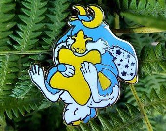 May include: A colorful enamel pin featuring a cartoon creature in blue, yellow, and white. The pin has a gold outline and is set against a backdrop of green fern leaves. The creature is holding a yellow star.