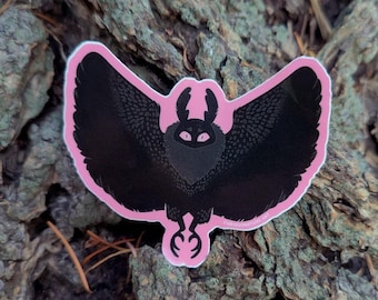 May include: A die-cut sticker featuring a stylized black moth with large, textured wings, prominent antennae, and glowing pink eyes. The moth is set against a solid pink background that follows the contour of its wings and body, creating a bold contrast.