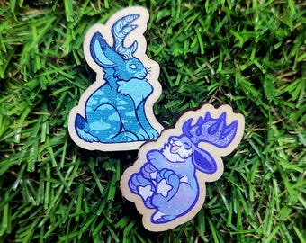 May include: Two wooden pins featuring mythical jackalopes. One pin shows a blue rabbit with antlers in a seated profile. The second pin depicts a purple rabbit with large antlers curled into a ball, displaying white star shapes on its paws.