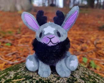 May include: A plush toy creature with gray fur, purple-lined ears, and black antlers and a fluffy neck. The toy has a white star on its forehead and is sitting on a mossy rock. The background features a blurred forest scene with autumn leaves.