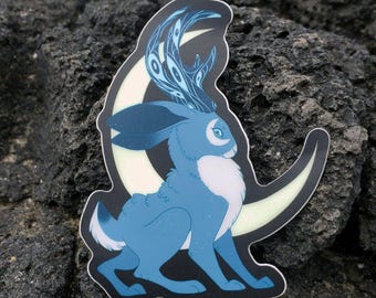 May include: A sticker featuring a blue jackalope with antlers and a crescent moon design. The jackalope has white fur on its chest and tail, with a starry pattern on its body. The moon is a light yellow color, and the sticker has a black border.