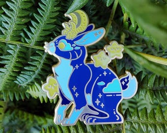 May include: A blue and light blue enamel pin in the shape of a rabbit with a crescent moon and star accents. The pin features a detailed design with a celestial theme, perfect for adding a touch of whimsy to any outfit or accessory.