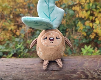May include: A plush toy resembling a Korok character from the Legend of Zelda series. The toy has a brown body with a smiling face, small arms and legs, and a large, light blue leaf on its head. It is sitting on a wooden rail.