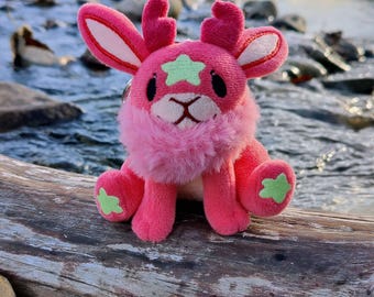 May include: A plush pink creature with a star on its head and green accents on its paws and head. The stuffed animal is sitting on a log near a body of water.