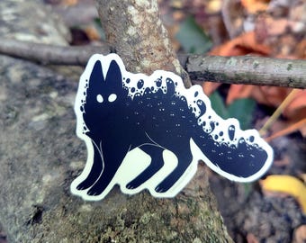 May include: A black sticker featuring a stylized fox silhouette with glowing white eyes and a tail adorned with small, ghostly figures. The sticker has a white border and is set against a natural, outdoor background with a tree branch and leaves.