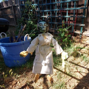 May include: A green-faced puppet with a white dress and yellow hands is hanging from strings in a backyard setting.