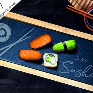 May include: A black slate serving tray with a wooden frame. The tray has a design of chopsticks and a circle with a line through it. The tray also has the text "Jack's Sushi" written on it. There are four pieces of sushi on the tray, including two pieces of salmon nigiri, one piece of avocado maki, and one piece of cucumber maki.