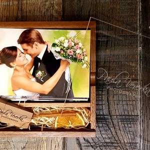May include: A wooden box with a clear acrylic lid, containing a wedding photograph of a couple embracing. The box is filled with dried orange slices and straw. The lid has the photographer's name, "Paula Venekla Photography", etched into it.