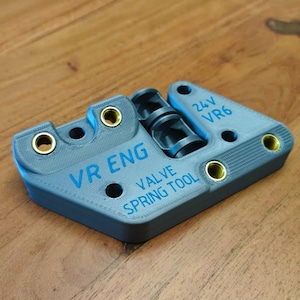 May include: A grey 3D-printed valve spring tool featuring blue text that reads VR ENG 24V VR6 VALVE SPRING TOOL. The component includes several circular openings with integrated brass-colored metal threaded inserts for mechanical fastening.
