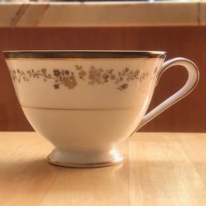 Puede incluir: Una taza de té blanca con un borde dorado y un patrón floral. La taza tiene un asa delicada y una base pequeña.