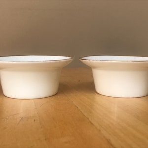 Puede incluir: Dos portavelas de cerámica blanca con borde dorado. Los portavelas están sobre una superficie de madera.