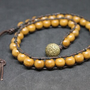 Peut inclure: Un cordon en cuir marron avec un collier de perles en bois. Le collier a une perle en métal doré avec une surface texturée.