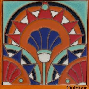 Op de afbeelding: Een vierkante keramische tegel met een geometrisch ontwerp met een gestileerde bloem met blauwe bloemblaadjes en een wit centrum. De bloem wordt omringd door een rood en oranje zonnestralenpatroon met zwarte accenten. De tegel heeft een turquoise rand en het woord "Outdoor" is afgedrukt in de rechterbenedenhoek.