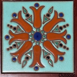May include: A turquoise blue ceramic tile with a symmetrical design of orange flower petals and blue and green accents. The tile has a brown wooden frame with the word "Outdoor" printed on the bottom.