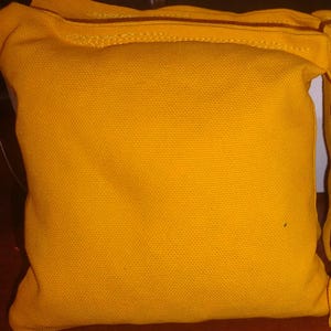 May include: Two square-shaped bean bags made of a solid, vibrant yellow fabric. Each bean bag features visible stitched seams along the edges, creating a defined border. The texture of the fabric appears woven and durable, with a consistent color.