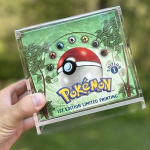 May include: A clear acrylic display case holding a Pokémon trading card. The card features a red and white Poké Ball, the Pokémon logo, and the text "1st Edition Limited Printing."