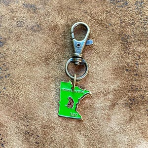 May include: A keychain with a metal clasp and a green enamel charm in the shape of Minnesota. The charm features a bear graphic and the text "NORTH STAR STATE" and "MN". The keychain is made of metal.