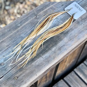 May include: A pair of long, feather earrings with a gold chain detail. The feathers are a mix of white, brown, and beige. The earrings are labeled "ASTALI" on a white card.