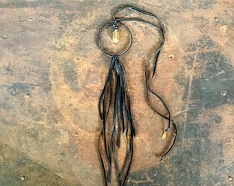Puede incluir: Un collar de cuero negro con un colgante circular dorado y una borla larga. El colgante tiene un cristal transparente en el centro.