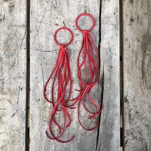 May include: A pair of red leather earrings with fringe tassels. The earrings are made of a thin, round leather strap with a small metal hoop at the top.