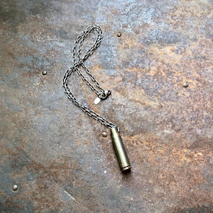 Puede incluir: Un collar de cadena de plata con un colgante de casquillo de bala. El colgante es de color dorado claro y tiene un acabado ligeramente deslustrado. El collar está sobre una superficie de metal oxidado.