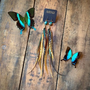 May include: A pair of long, colorful feather earrings with turquoise beads. The earrings are handmade in the USA and are displayed on a wooden surface with a butterfly.