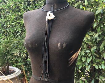May include: A black beaded necklace with a white stone pendant and a long black leather tassel. The necklace is displayed on a black mannequin against a backdrop of green foliage.