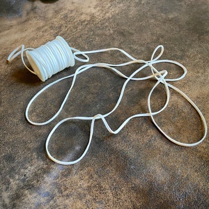 Puede incluir: Un carrete de cordón elástico blanco, con una parte del cordón desenrollada y sobre una superficie marrón.
