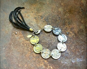 May include: A silver bracelet with a black leather tassel. The bracelet is made of a chain with a series of round silver and gold coins attached. The coins have a raised design.