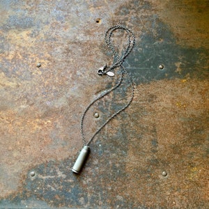 May include: A silver chain necklace with a silver bullet pendant. The necklace is lying on a rusty metal surface.