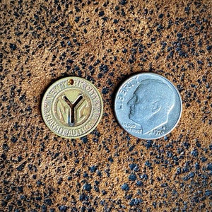 May include: A vintage New York City Transit Authority token with the NYC logo, next to a 1976 silver dime. The token is gold-colored and circular, with text around the edge. The dime shows a profile of a person.