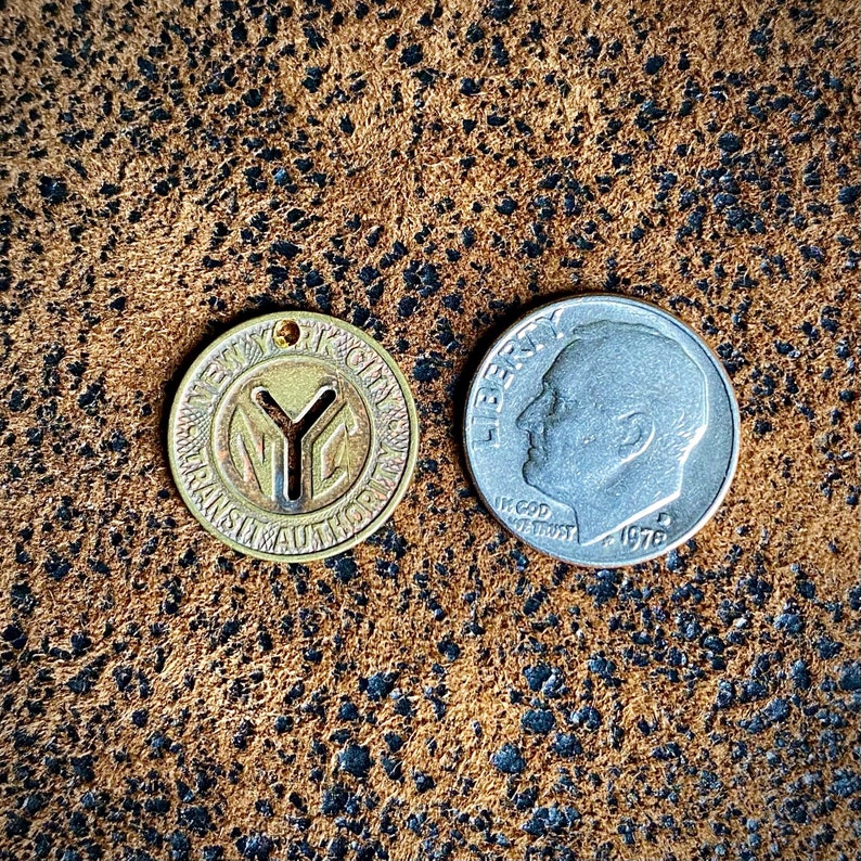 May include: A vintage New York City Transit Authority token with the NYC logo, next to a 1976 silver dime. The token is gold-colored and circular, with text around the edge. The dime shows a profile of a person.