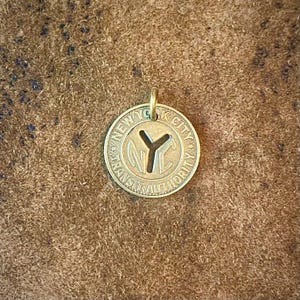 May include: A circular brass pendant featuring the text "NEW YORK CITY TRANSIT AUTHORITY" around the edge. The center has a cutout of the letters "NYC" and a "Y" shape. The pendant has a small loop at the top for a chain.